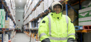 A female warehouse operator wearing a Voice Picking headset and high-visability clothing within an aisle of pallet racking.
